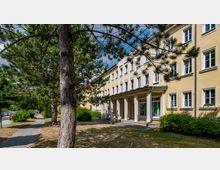 Gelbes, mehrstöckiges Gebäude im klassischen Stil mit weißen Fensterrahmen und Säulen am Eingangsbereich. Im Vordergrund stehen Bäume, ein gepflasterter Gehweg führt durch eine Grünfläche.