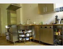 Moderne Küche mit Edelstahlmöbeln, einschließlich eines Herds mit Dunstabzugshaube, einer Spüle und einer Geschirrspülmaschine. Die Arbeitsflächen bestehen aus Holz, die Rückwand ist mit weißen Fliesen verkleidet, und offene Regale sowie geschlossene Schränke bieten Stauraum für Küchenutensilien und Lebensmittel.