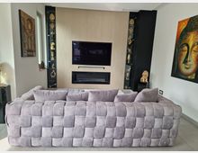 Wohnzimmer mit modernem Design: Eine graue, gepolsterte Couch steht vor einer Wand mit hellem Holz und integriertem Flachbildfernseher sowie einem Kamin. Auf beiden Seiten der Wand befinden sich offene Regale mit goldfarbenen Dekorobjekten. An den Wänden hängen buddhistische Kunstwerke, darunter ein großes Buddha-Gemälde.