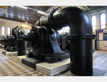 Industrieller Innenraum mit großen schwarzen Maschinen aus Metall, vermutlich Pumpen oder Turbinen, auf gefliesten Sockeln. Die Umgebung zeigt hohe Fenster mit Rundbögen und helle Wände in einem historischen Gebäude.