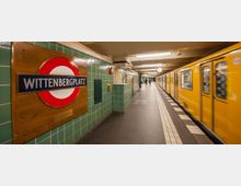 U-Bahnhof Wittenbergplatz in Berlin mit grün gefliesten Wänden, einem großen Holzschild mit Stationsnamen und einem gelben U-Bahnzug auf der rechten Seite, der an einem beleuchteten Bahnsteig hält. Im Hintergrund sind eine runde Bahnhofsuhr und Passanten zu sehen.