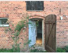 Backsteingebäude mit einem offenen Holztor und einer halb geöffneten weißen Tür, die in einen Raum mit Fliesenboden führt. Rechts oben ist eine Außenlampe montiert, während links eine kleine Pflanze an der Fassade entlangwächst.