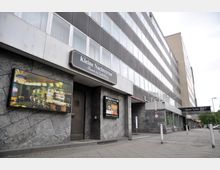 Ansicht einer Fassade mit dem Schriftzug „Kleine Nachtrevue – Theater der Enthüllung“ über einem Eingang, flankiert von Werbetafeln. Das Gebäude ist mehrstöckig mit dunklen, quadratischen Fenstern und einem grauen, strukturieren Wandbelag, daneben ein Schild des benachbarten „Hotel Sylter Hof“.