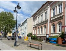 Straßenszene mit einer Reihe historischer Gebäude auf der rechten Seite, geprägt von verzierten Fassaden und hohen Fenstern. Auf der linken Seite stehen Laternen und ein Baum, während geparkte Autos und ein gepflasterter Gehweg das Stadtbild ergänzen.