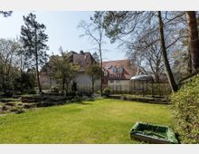 Weitläufiger Garten mit Rasenfläche, Bäumen und Sträuchern, im Hintergrund ein großes Wohnhaus mit Satteldach und Dachgauben. Rechts im Bild steht ein metallener Pavillon, umgeben von Bäumen und Hecken.