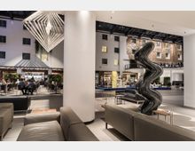 Ein moderner Hotel-Lobbybereich mit eleganten Ledersofas, einem geschwungenen schwarzen Kunstwerk im Vordergrund und einer großen, offenen Sitzfläche mit Tischen im Hintergrund. Die Architektur zeigt hohe weiße Säulen, eine verglaste Decke und eine Wand mit dekorativen Mosaikfliesen.