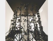 Ein Raum mit weiß gestrichenen Wänden und schwarzer Decke, in dem mehrere Stative mit Kameras und Beleuchtungssystemen dicht positioniert sind, verbunden durch zahlreiche Kabel. Im Hintergrund sind Fenster zu sehen, die Tageslicht hereinlassen.