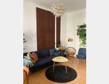 Wohnzimmer mit moderner Einrichtung: Eine dunkelblaue Couch, dekoriert mit Kissen, steht vor einer Wand mit vertikalen Holzpaneelen. Eine runde Deckenlampe aus Glasbubbles hängt zentral über einem beigefarbenen Couchtisch, der auf einem schwarzen Teppich steht. Pflanzen, ein goldfarbener Bogenstrahler, ein Sessel, sowie ein Holzstuhl runden das minimalistische Design ab.