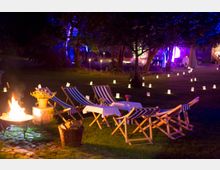 Nachtszene in einem Garten mit Liegestühlen und kleinen Tischen auf einer Wiese, umgeben von Kerzenlichtern in Papierlaternen. Im Vordergrund eine Feuerstelle und ein Korb mit Holzscheiten, im Hintergrund beleuchtete Bäume in Blau- und Violetttönen.