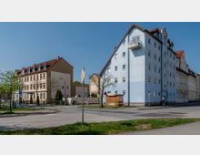 Straßenecke mit mehrstöckigen Wohngebäuden, darunter ein weiß-blaues Haus und ein beigefarbenes Gebäude mit rotem Satteldach. Im Vordergrund eine gepflegte Straße mit Verkehrszeichen sowie ein kleiner bepflanzter Grünstreifen.