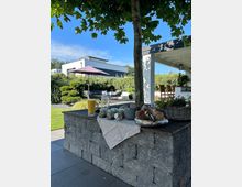 Überdachte Terrasse in einem modernen Garten mit einer Sitzlounge, Sonnenschirm und gepflegten Hecken im Hintergrund. Im Vordergrund steht ein gemauerter Außenbereich mit Ablagefläche unter einem Baum, darauf Gläser und Tabletts mit Frühstücks- oder Snackutensilien.