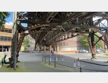 Eine stählerne Hochbahntrasse, die mit rot-weißen Markierungen versehen ist, überspannt eine Straße neben Backsteingebäuden und einem modernen Parkhaus. Im Vordergrund stehen ein Roller, Poller und Straßenpfosten, während im Hintergrund geparkte Autos und Fußgänger erkennbar sind.