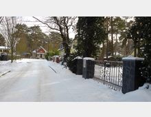 Verschneite Wohnstraße mit freistehenden Häusern und Vorgärten, umgeben von hohen Bäumen. Im Vordergrund ein schmiedeeisernes Tor mit steinernen Pfosten, im Hintergrund ein rotes Haus am Ende der Straße.