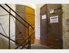 Eine Treppe mit einem Metallgeländer führt zu zwei großen, älteren Metalltüren in einem hell gestrichenen Backsteinflur. An den Türen sind Schilder angebracht, darunter eines mit der Aufschrift "Kindertanz".