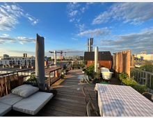 Dachterrasse mit Holzboden und Sitzmöbeln, darunter ein Tisch mit gestreifter Tischdecke und ein gepolsterter Liegestuhl. Die Terrasse ist mit Pflanzen, Bambusabtrennungen und einem kleinen Holzsaunahaus dekoriert. Im Hintergrund sind Wohngebäude, ein Kran und ein klarer Himmel zu sehen.