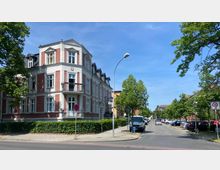 Straßenszene mit einem dreigeschossigen Wohngebäude im Gründerzeitstil aus rotem Backstein und weißen Fassadenelementen an einer Straßenecke. Rechts führt eine gepflasterte Nebenstraße mit parkenden Autos und Bäumen weiter, während links eine breitere Straße mit Gehweg verläuft.