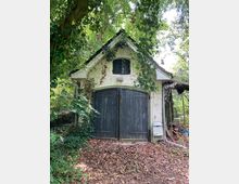 Kleines Gebäude mit weiß verputzter Fassade und schwarzem Giebeldach, umgeben von dichtem Wald. Das Gebäude hat große, doppelflügelige Holztüren und ist teilweise von Efeu bewachsen; Laub bedeckt den Boden davor.