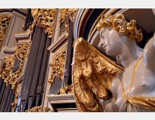 Nahaufnahme eines prunkvollen Barock-Orgelgehäuses mit vergoldeten Verzierungen und einem detailreichen Engelsornament mit goldenen Flügeln. Im Hintergrund sind große Pfeifen und filigrane Ornamentik zu sehen.