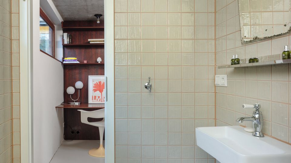 Small tiled bathroom with light square wall tiles, a wall-mounted white sink and chrome faucet beneath a shelf and mirror. An open doorway leads to an adjacent room with built-in dark wood shelving, a desk area, and a window.