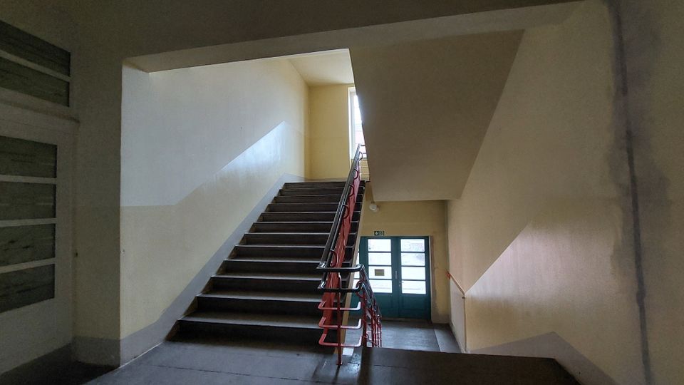Treppenhaus in einem Gebäude mit gelb gestrichenen Wänden und einem Handlauf in Rot. Eine Treppe führt hinauf zu einem Fenster, das Tageslicht hineinlässt, und hinunter zu einer Tür mit Glaselementen.
