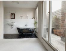 Minimalistisches Badezimmer mit freistehender, dunkler Badewanne auf schwarzen Fliesen, weiß gefliester Rückwand mit einem kleinen Waschbecken und Spiegel. Rechts befinden sich große Fenster, die einen Blick auf eine Terrasse mit Topfpflanzen bieten.
