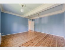 Leerer Innenraum mit hellblauen Wänden und breiten, lackierten Holzdielen. Weiße Stuckdecken mit Querbalken und zwei runden Deckenleuchten sowie eine offene Tür zu einem angrenzenden Flur.