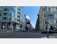 Straßenszene in Berlin an der Kreuzung Zimmerstraße, mit Blick auf den Checkpoint Charlie. Links ein modernes Gebäude mit Glasfassade, darunter eine KFC-Filiale, rechts historische Gebäude mit klassischer Architektur; mittig ein großes Plakat mit dem Bild eines Soldaten.