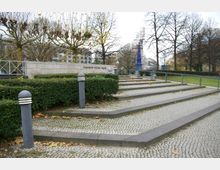 Städtischer Platz mit gepflasterten Stufen und umliegender Grünanlage, in der Mitte eine Hecke und ein Schild mit der Aufschrift "Theodor-Heuss-Platz". Im Hintergrund sind kahle Bäume, Wohngebäude und eine blaue Säule zu sehen.