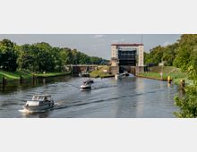 Flusslandschaft mit einer Schleuse im Hintergrund, umgeben von grünen Uferbereichen und Bäumen; mehrere Boote und kleine Schiffe fahren in der Wasserstraße auf die Schleuse zu.