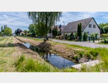 Eine ländliche Wohnsiedlung mit freistehenden Häusern und Vorgärten, flankiert von einem schmalen Kanal, der durch eine grasbewachsene Fläche verläuft. Im Hintergrund sind Bäume und weitere Gebäude zu sehen, während eine Straße am rechten Ufer entlangführt.