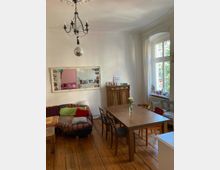 Wohnzimmer mit Holzboden, einem langen Holztisch und Stühlen. Links steht ein bunt gemustertes Sofa, darüber ein großer Wandspiegel, und an der Decke hängt ein Kronleuchter sowie eine Discokugel. Rechts befinden sich ein Fenster mit weißem Vorhang und ein hölzernes Sideboard.