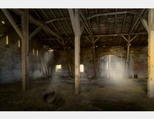 Großer, leerer Innenraum einer alten Scheune mit Holzstützbalken, offenen Dachbalken und Wänden aus Ziegelstein. Durch kleine Fenster und Risse in der Struktur fällt diffuses Licht in den staubigen Raum, der teilweise mit Stroh bedeckt ist.