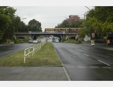 Stadtstraße mit geteilter Fahrbahn und mittlerem Grünstreifen, im Hintergrund eine Brücke mit einer fahrenden S-Bahn. Umgeben von Bäumen und Gebäuden, mit gelegentlich parkenden Autos und Straßenschildern.