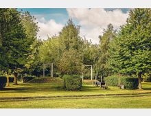 Ein grüner Park mit Rasenflächen, hohen Bäumen und Büschen. Im Vordergrund verlaufen schmale Gleise, daneben stehen Bänke und ein Spielbereich mit Holzrahmenstrukturen.