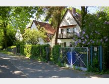 Wohnstraße mit einem kleinen Einfamilienhaus mit Satteldach, weißer Fassade und Balkon. Der Vorgarten ist dicht bepflanzt mit Bäumen, Sträuchern und blühendem Flieder, während ein Metallzaun mit einem Tor den Garten von der Gehwegseite abgrenzt.