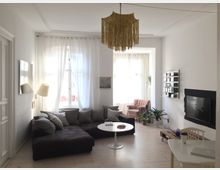 Helles Wohnzimmer mit einem großen braunen Ecksofa, weißen Vorhängen vor zwei Fenstern, einer goldenen Lampe an der Decke und einem weißen runden Couchtisch. An der Wand hängt ein Flachbildfernseher, daneben stehen Regale mit Büchern und Zimmerpflanzen; ein rosa Sessel und ein Schreibtisch sind ebenfalls zu sehen.