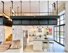 Moderne Wohnküche in einem offenen Loft mit Galerieebene. Sichtbare weiße Backsteinwände, eine großzügige Kücheninsel mit Hockern, farbenfrohe Kunstwerke an den Wänden und große Fenster mit schwarzem Metallrahmen schaffen ein helles, industrielles Ambiente.