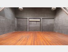 Ein leerer, rechteckiger Raum mit dunkelgrauen Wänden und einem polierten Holzboden. An der Rückwand befindet sich eine horizontale Halterung mit Farbrollen, und von der Decke hängen silberne Lampenschirme.