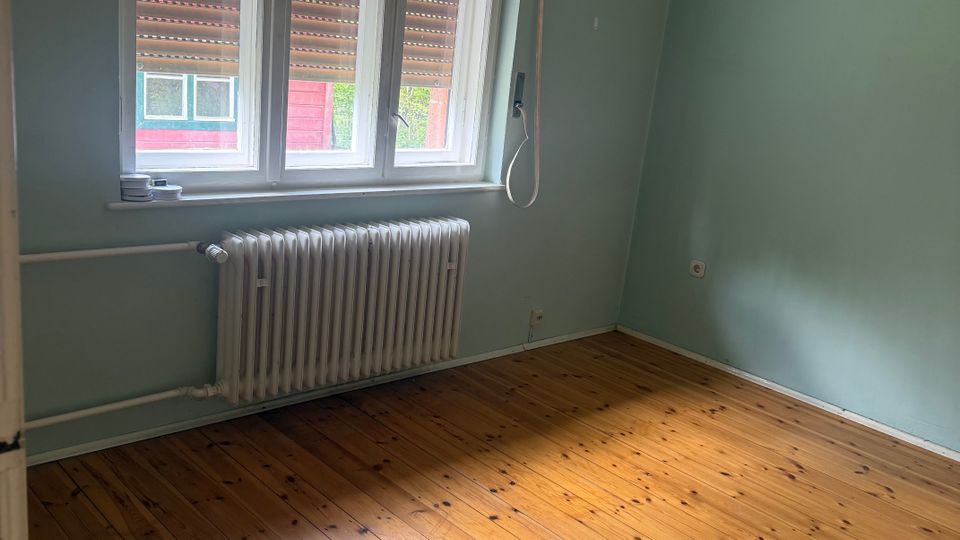Leeres Zimmer mit hellblauen Wänden und holzfarbenem Dielenboden, beleuchtet durch ein großes dreiteiliges Fenster. Unter dem Fenster befindet sich ein weißer Heizkörper; außen sind Rollläden teilweise heruntergelassen.
