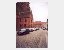 Straßenszene mit Kopfsteinpflaster, links eine Backsteinkirche mit spitzem Dach, dahinter weitere historische Gebäude. Mehrere Autos sind am Straßenrand geparkt.