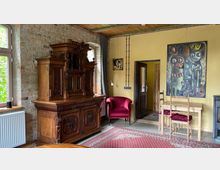 Wohnraum mit rustikalem Stil: An der gelben Wand stehen ein massiver, verzierter Holzschrank und ein roter Polstersessel. Daneben ein kleiner Holztisch mit zwei Stühlen, ein modernes Kunstwerk hängt an der Wand. Die Wand besteht teils aus Ziegelsteinen, und der Boden ist mit einem roten Teppich bedeckt. Ein großes Fenster links lässt Licht herein.