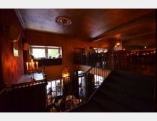 Zweistöckiges Café mit gemütlicher Beleuchtung, dekorativen Wandleuchten und einem Kronleuchter. Die obere Etage verfügt über Sitzplätze mit Holzstühlen und Geländer, während die untere Etage einen Blick nach draußen durch große Fenster bietet.