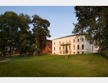 Historisches Gebäudeensemble mit einer weißen Villa und einem angrenzenden roten Ziegelgebäude, umgeben von einer weitläufigen Wiese und großen Bäumen, bei Sonnenuntergang.