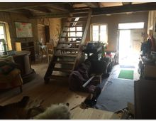 Rustikaler Innenraum eines Hauses mit unverputzten Lehmwänden, Holzboden und einer Holztreppe, die zu einem offenen Zwischengeschoss führt. Im Raum befinden sich verschiedene Möbel, darunter ein runder Tisch, Stühle, Regale und ein Sofa, mit einer offenen Tür, die nach außen führt und für Tageslicht sorgt.