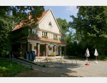 Zweistöckiges Gebäude mit holzverkleideter Fassade und einem roten Ziegeldach, umgeben von Bäumen. Im Vordergrund ein Außenbereich mit zusammengeklappten Stühlen, Tischen und Sonnenschirmen auf einem Kiesplatz.