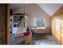 Helles Arbeitszimmer mit Holzwänden und schräger Decke. Der Raum enthält einen Schreibtisch vor einem Fenster mit Blick nach draußen, einen modernen Bürostuhl, ein Regal mit Büchern und Dekorationsgegenständen sowie eine Hängesitzschaukel mit rotem Kissen. Rechts befindet sich eine Wand mit eingebauten Holzschränken.