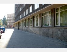 Straßenecke mit einem dunkel verputzten Gebäude auf der rechten Seite, dessen Fassade durch große rechteckige Fenster gegliedert ist, und einem historischen Gebäude im Hintergrund links. Der Gehweg besteht aus grauen Betonplatten, daneben parken einige Autos.