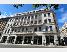 Straßenszene mit einem mehrstöckigen Gebäude in moderner klassizistischer Architektur, gekennzeichnet durch helle Fassaden mit symmetrisch angeordneten Fenstern, Balkonen und Säulen. Im Erdgeschoss befinden sich Schaufenster, davor stehen Topfpflanzen, und ein Bürgersteig verläuft entlang der Straße.