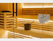 Innenansicht einer modernen Sauna mit hellen Holzlamellen, mehrstufigen Sitzbänken und warmem indirektem Licht. Links steht ein Saunaofen mit Steinen, davor ein Aufgusseimer mit Kelle; an der Glaswand hängt ein zusammengerolltes Handtuch.