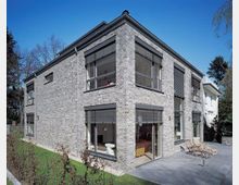 Eckansicht eines modernen Wohnhauses mit zweistöckiger Backsteinfassade, großen Fenstern und grauen Lamellenrollos. Der Außenbereich umfasst eine Terrasse mit Liegestühlen sowie angrenzende Grünflächen.